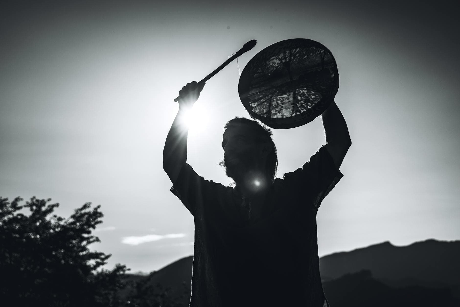 A dramatic black-and-white silhouette of a shaman holding a drum against the mountainous backdrop of Lombardia, Italy.